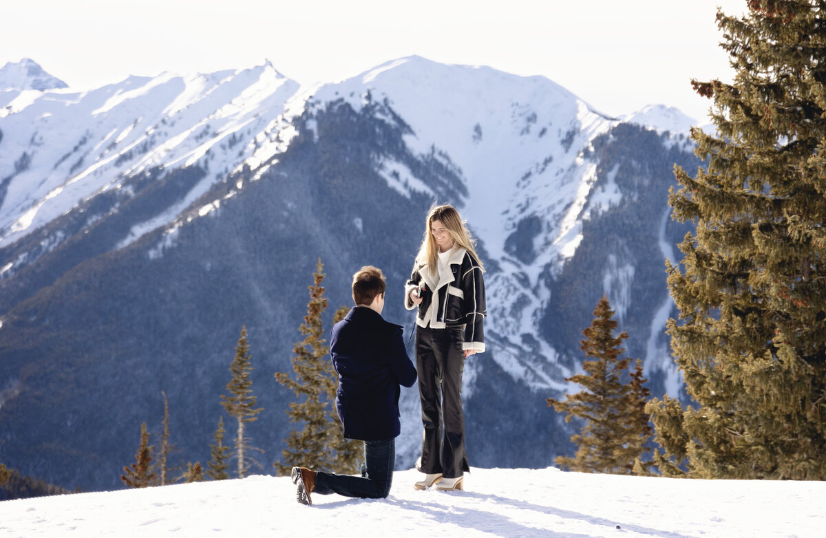 a young man is on his knees asking his girlfriend to marry him on top of aspen mountain with the most beautiful scenery