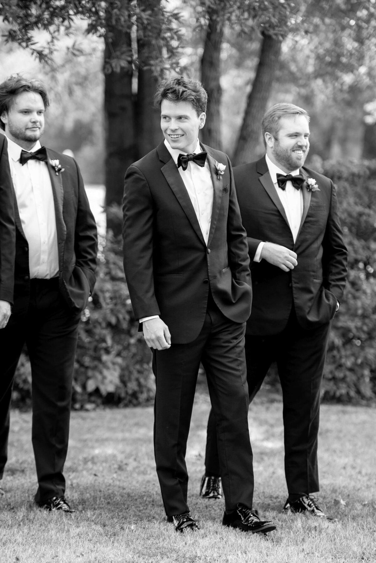 Three men in tuxedos stand outside on grass, smiling and chatting. They are wearing bow ties and boutonnières. Trees and bushes are in the background. The image is in black and white.