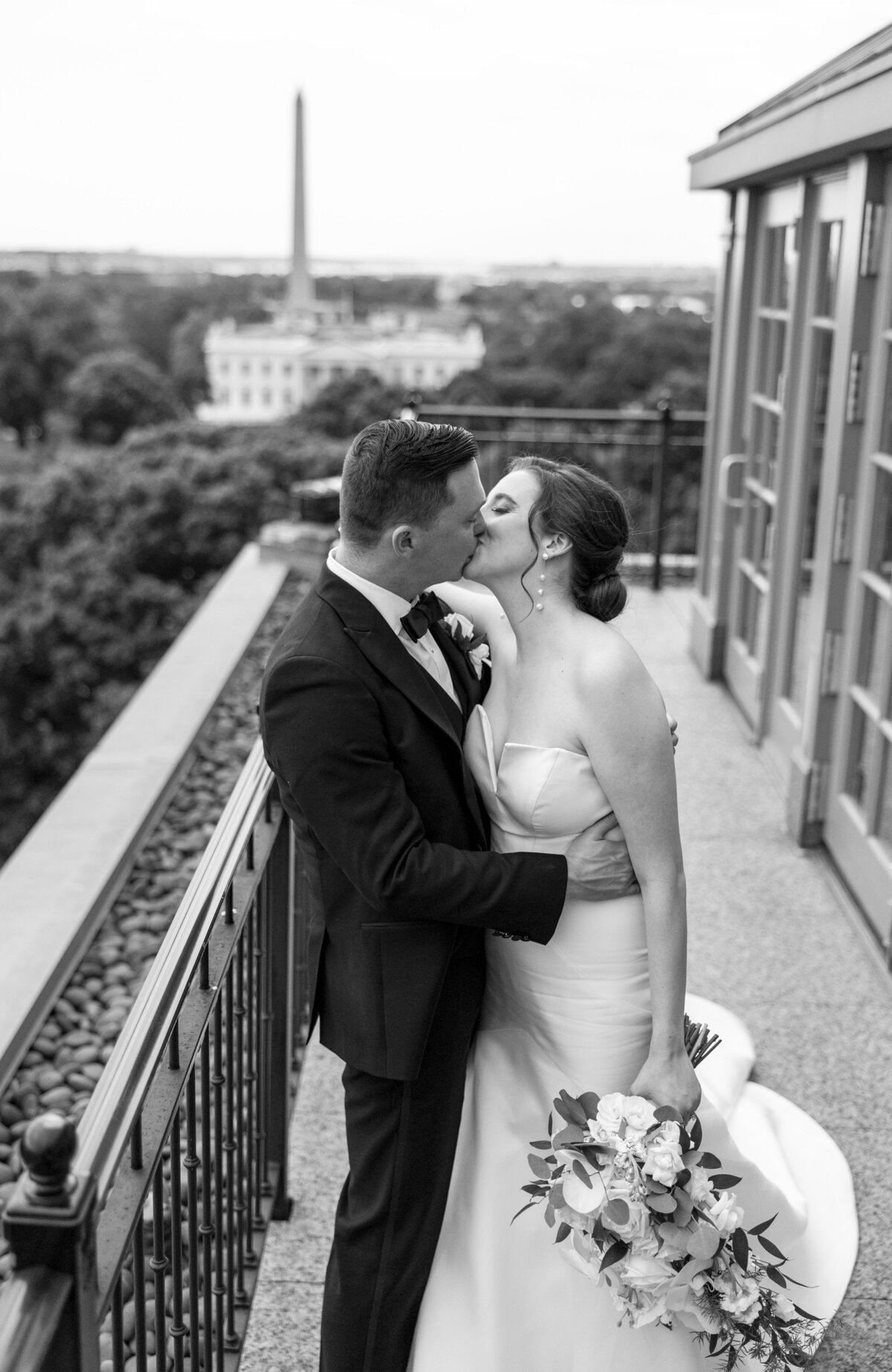 A bride and groom share a kiss on a balcony overlooking a cityscape with a tall monument in the background. The bride holds a bouquet and wears a strapless gown, while the groom is in a suit. Black and white photo.