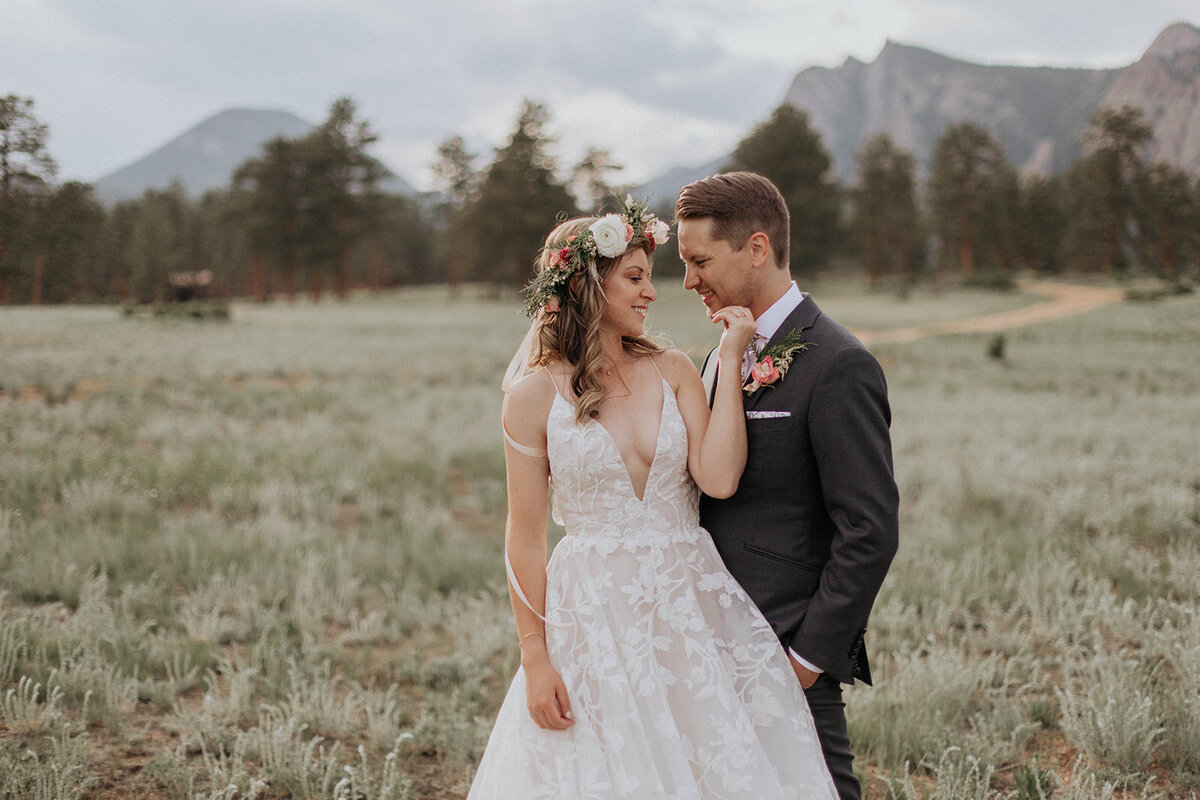 A bride and groom share a loving moment in a scenic mountain meadow, with the bride wearing a floral crown and a flowing lace gown.