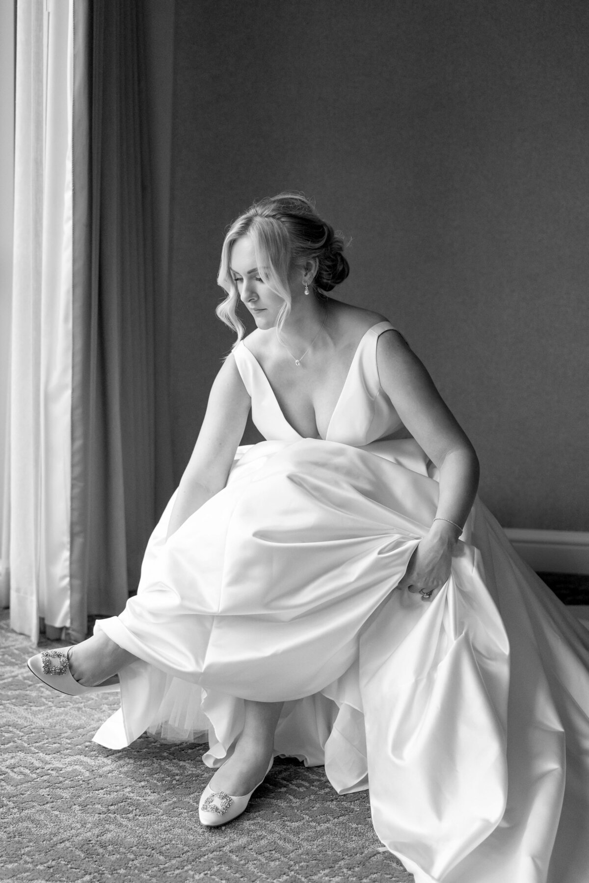 A black and white image of a woman in a wedding dress sitting near a window. She is adjusting her shoe, with her hair styled in an elegant updo and the dress featuring a flowing skirt and a deep V-neckline.