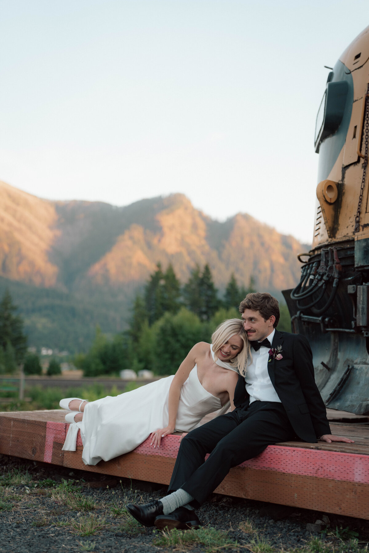 Bride resting on groom’s shoulder sitting by vintage train with mountain backdrop