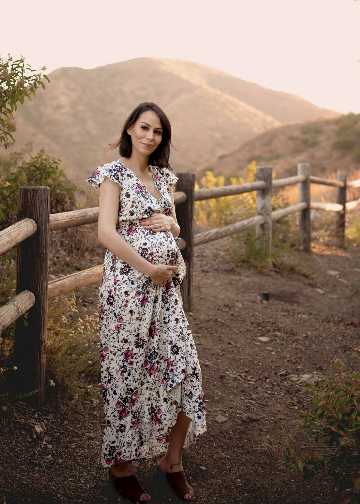 Expectant Mom in long, floral dress smiling at the camera with the Elsinore mountains in the distance.