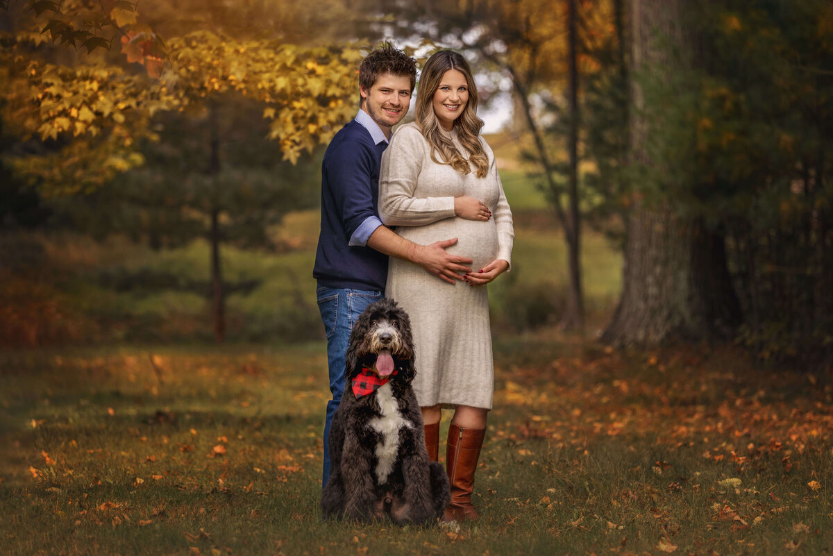 Expecting Couple Standing In Fall Leaves With Pet Dog