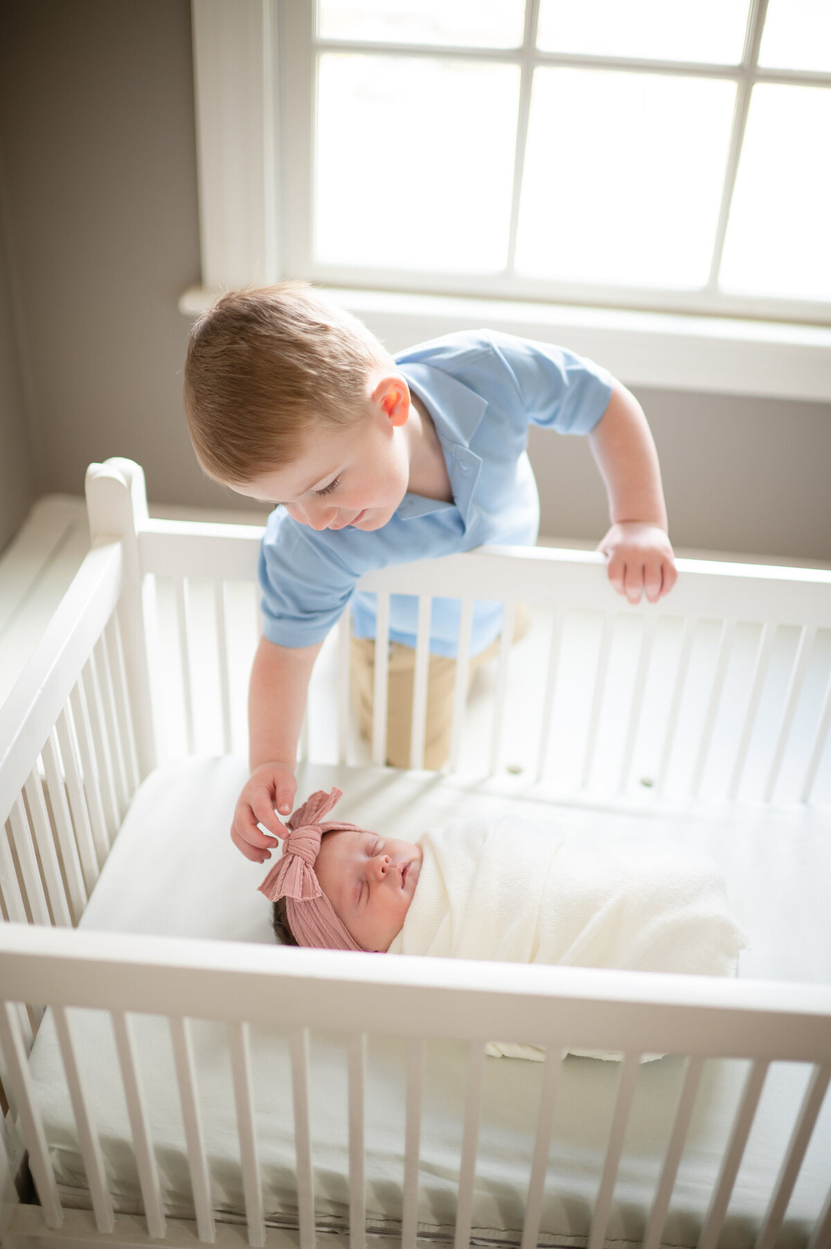 Toddler boy reaches into crib to pat his newborn sibling's head.