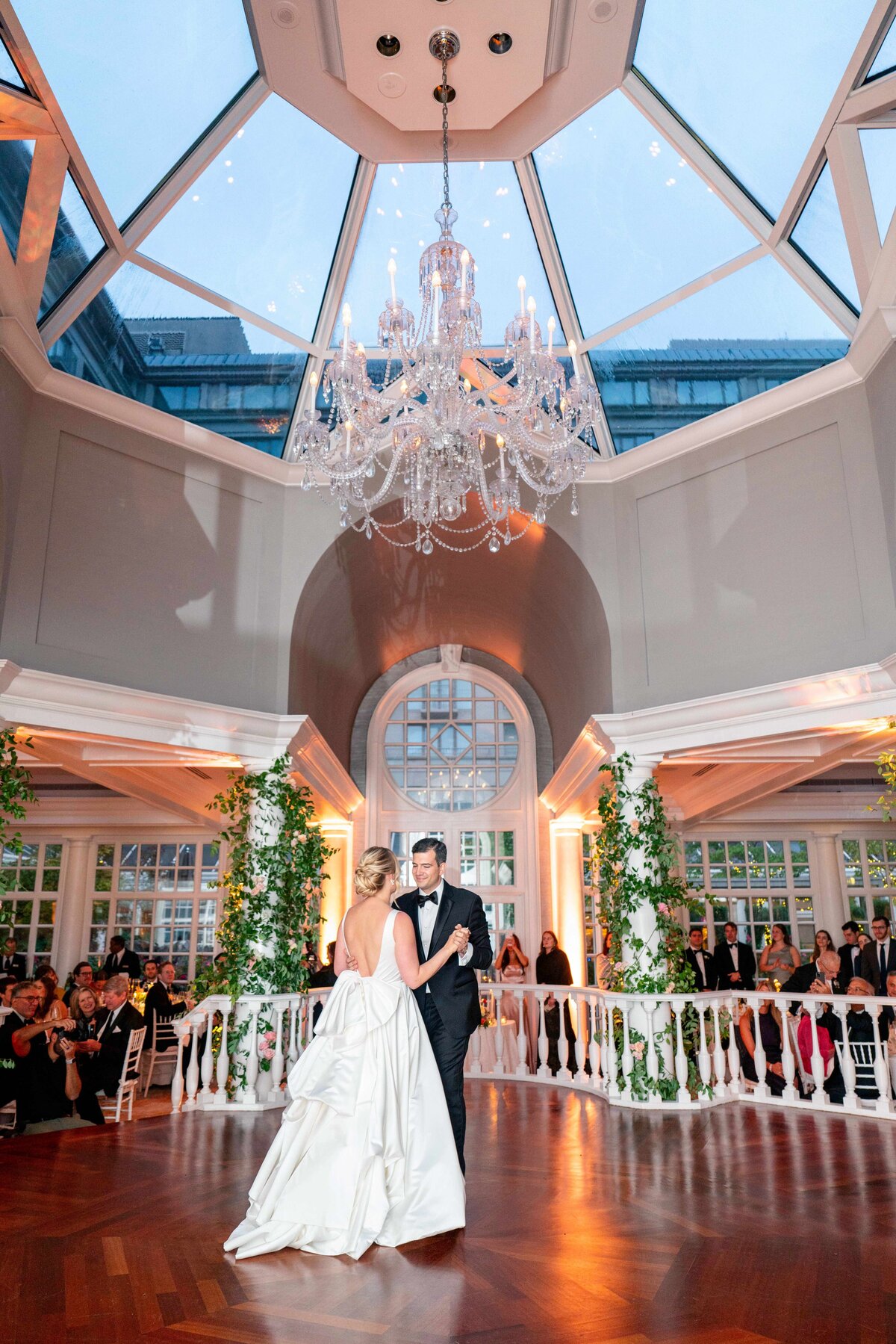 A couple is dancing under a grand chandelier in an elegant ballroom with a high glass ceiling. The bride is in a white gown, and the groom is in a black suit. Guests are seated, watching the dance. Greenery adorns the hall.
