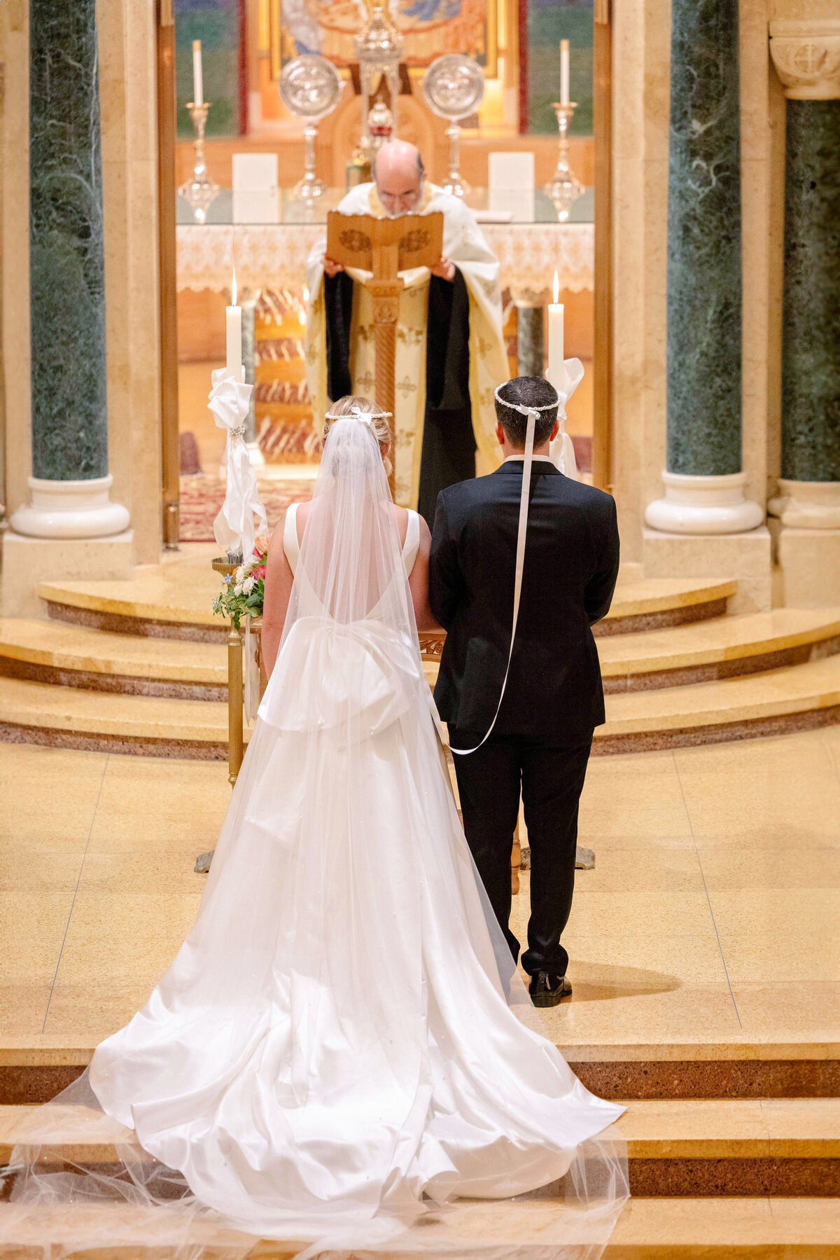 A bride in a white gown and veil stands next to a groom in a black suit, both facing an altar inside a church. A priest, holding a book, stands at the altar, performing a ceremony. Candles adorn the altar, and pillars flank the scene.
