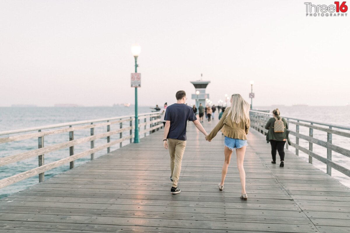 Engaged couple walk away on the Seal Beach Pier holding hands