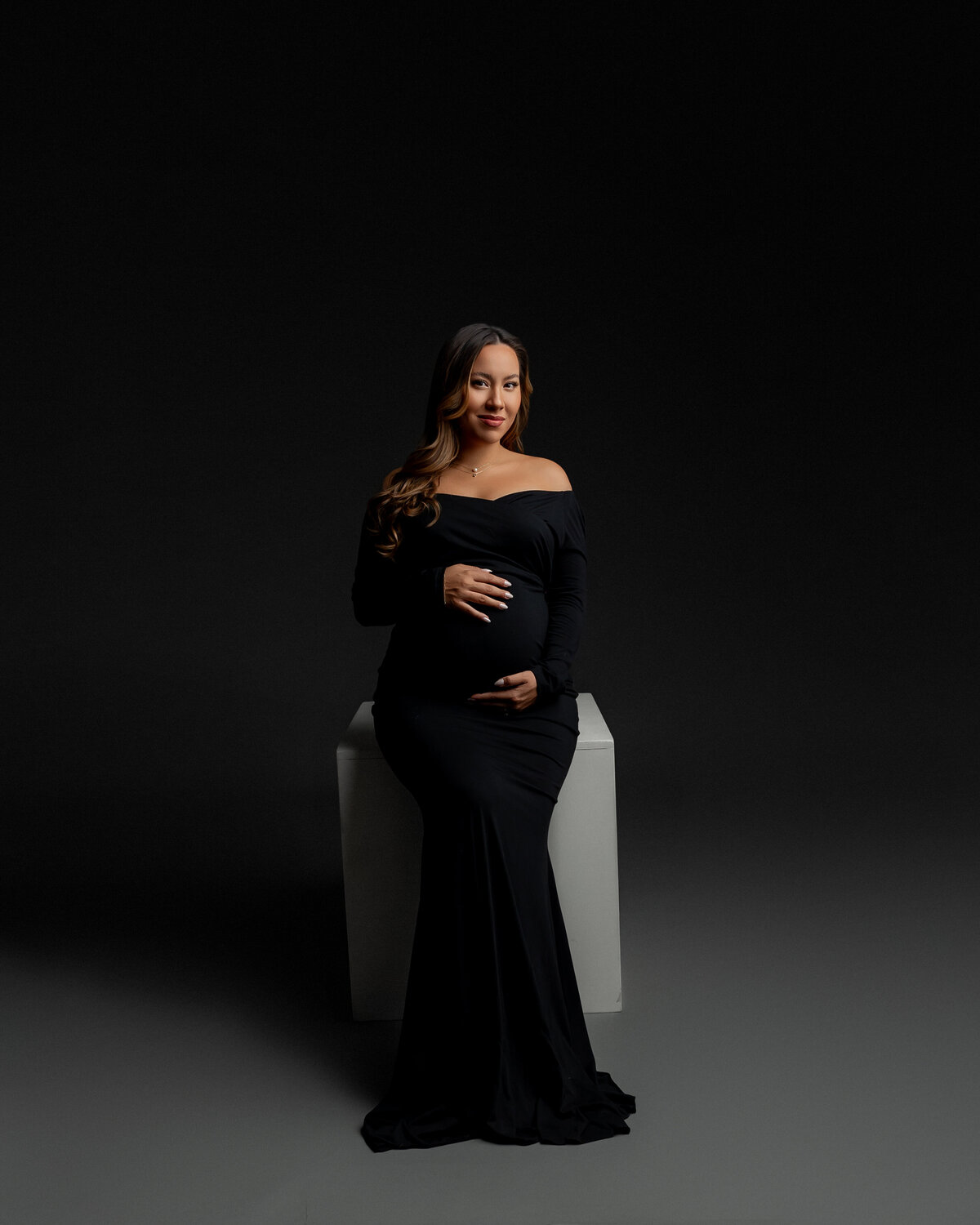 Sitting gracefully in a black dress, she gently holds her belly bump, capturing a serene moment of motherhood. Captured by Inland Empire photographer Kaitlyn Dawn Photography.