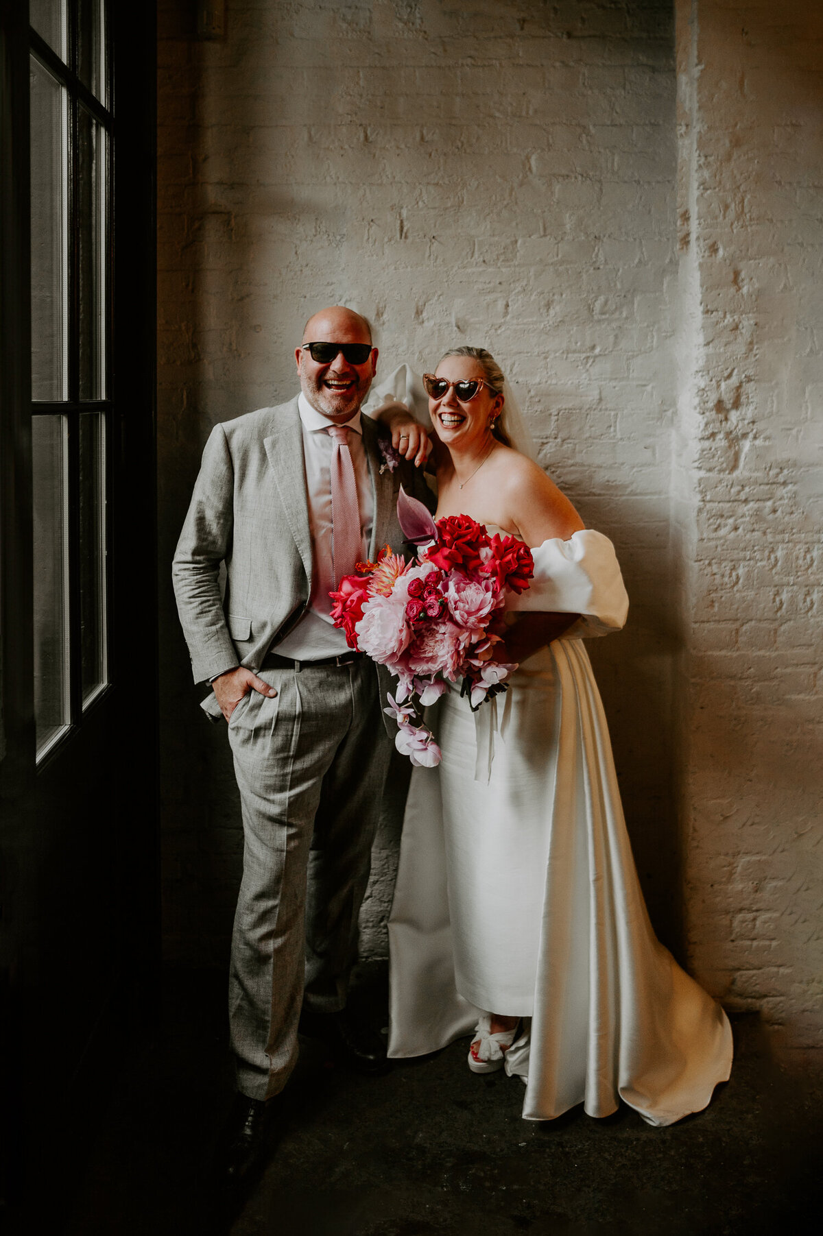 A bride and groom stand in the doorway of 100 Barrington in London wearing sunglasses, the bride is holding a pink bouquet and the groom's tie matches the flowers.