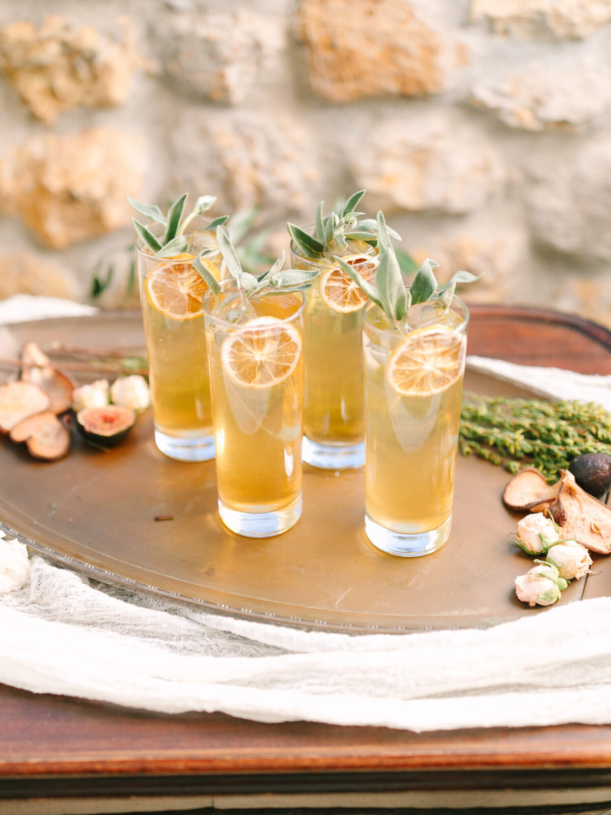 Four tall glasses of cocktail garnished with sage and dried lemon slices sit on a rustic tray.