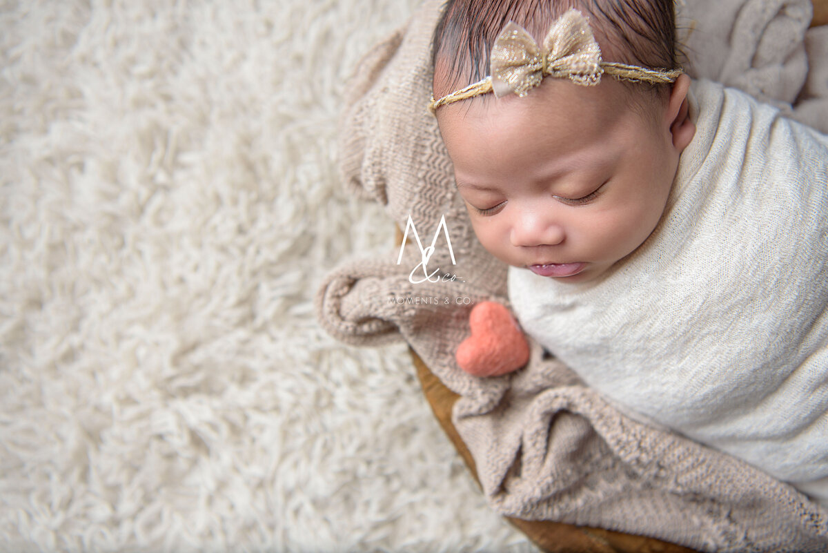 Newborn-Girl-Wrapped-in-tan-with-headband-bow-and-pink-heart-in-bowl-on-fur