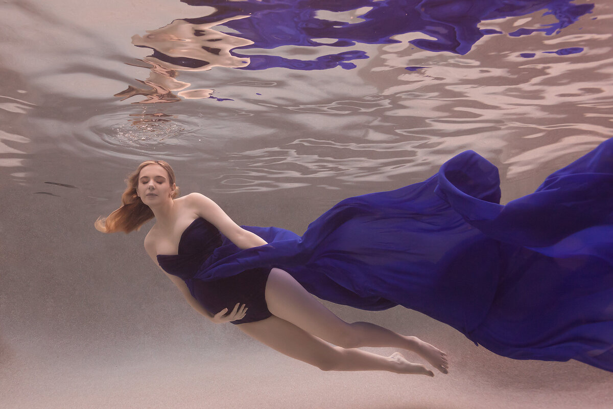 An ethereal underwater maternity photo captured by a skilled Dallas underwater maternity photographer. The woman is elegantly floating in the water, her rich blue gown flowing gracefully around her, creating a dreamlike scene. Her relaxed posture and closed eyes convey a sense of peace and calm, perfectly embodying the beauty of underwater maternity photography.