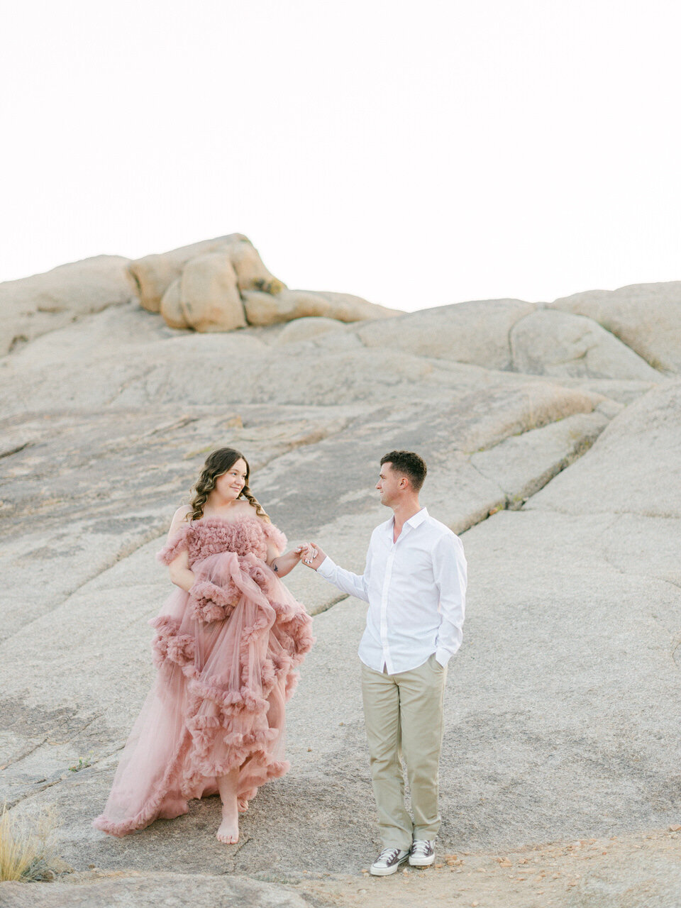 A couple exploring the desert boulders in Joshua Tree, CA, with the woman wearing a flowing dusty-pink gown, captured in a refined editorial style by Elizabeth Kane Photography—serving Philadelphia, New York, and DC.