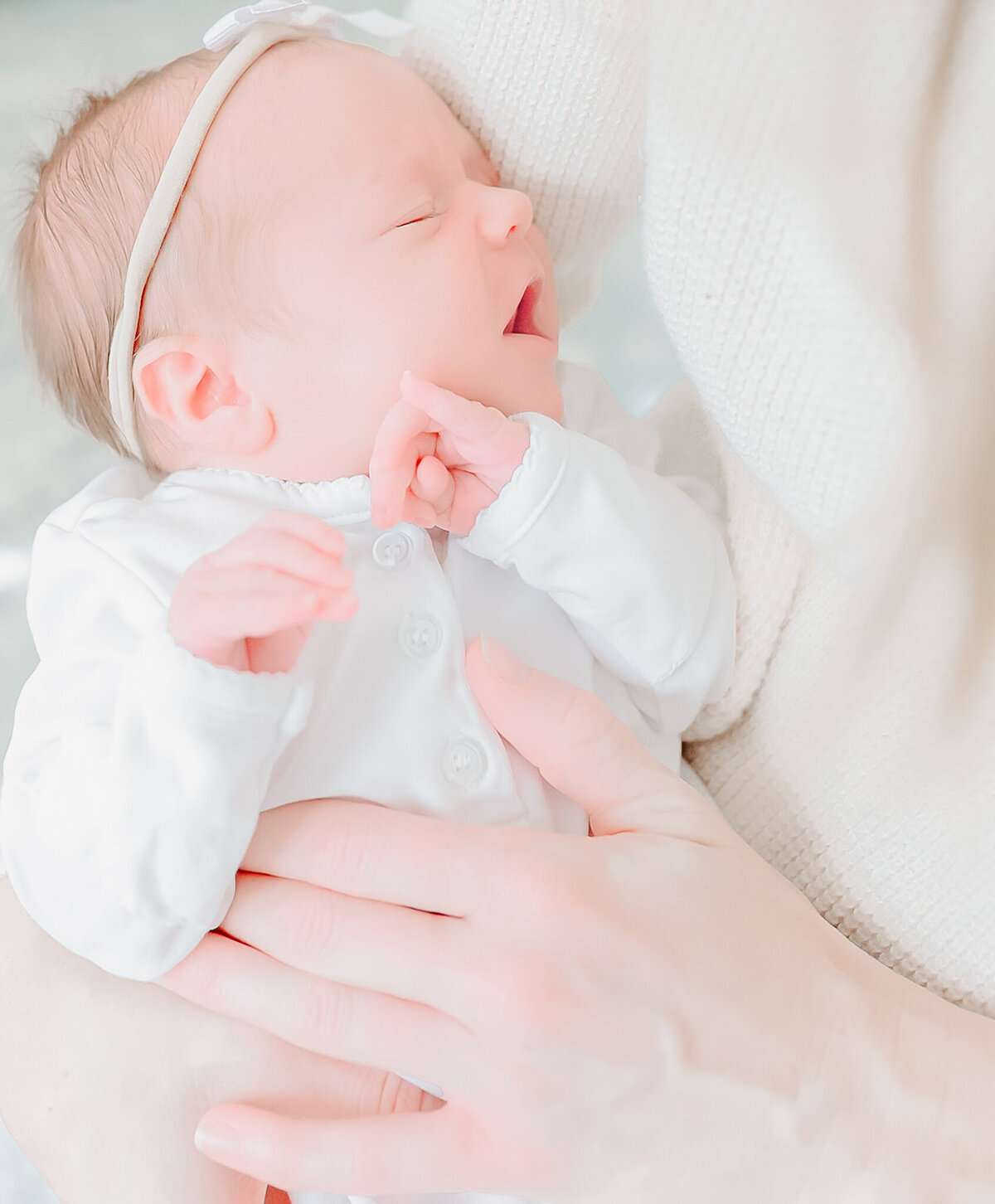 A newborn baby sleeps peacefully in her mother’s arms, dressed in a soft white outfit with a delicate headband. This light and airy portrait captures the tenderness of early motherhood, reflecting the warm and natural style of a Birmingham newborn photographer.