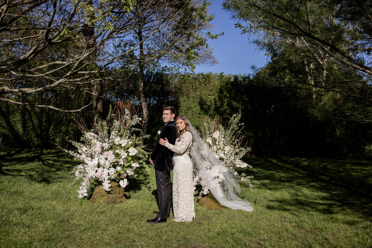 Bride and groom pose back-to-back for an editorial-style wedding portrait surrounded by sculptural floral arrangements in a private Hamptons garden setting.