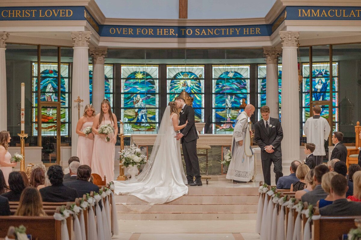 A Catholic wedding ceremony at St. Joseph’s Catholic Church in Jacksonville, Florida, as the bride and groom share their first kiss at the altar. The bride wears a long cathedral veil and white gown, surrounded by bridesmaids in blush pink dresses and groomsmen in black suits. Behind them, stained glass windows glow with rich blue and green hues. Phavy Photography, Wedding Photographer in Jacksonville