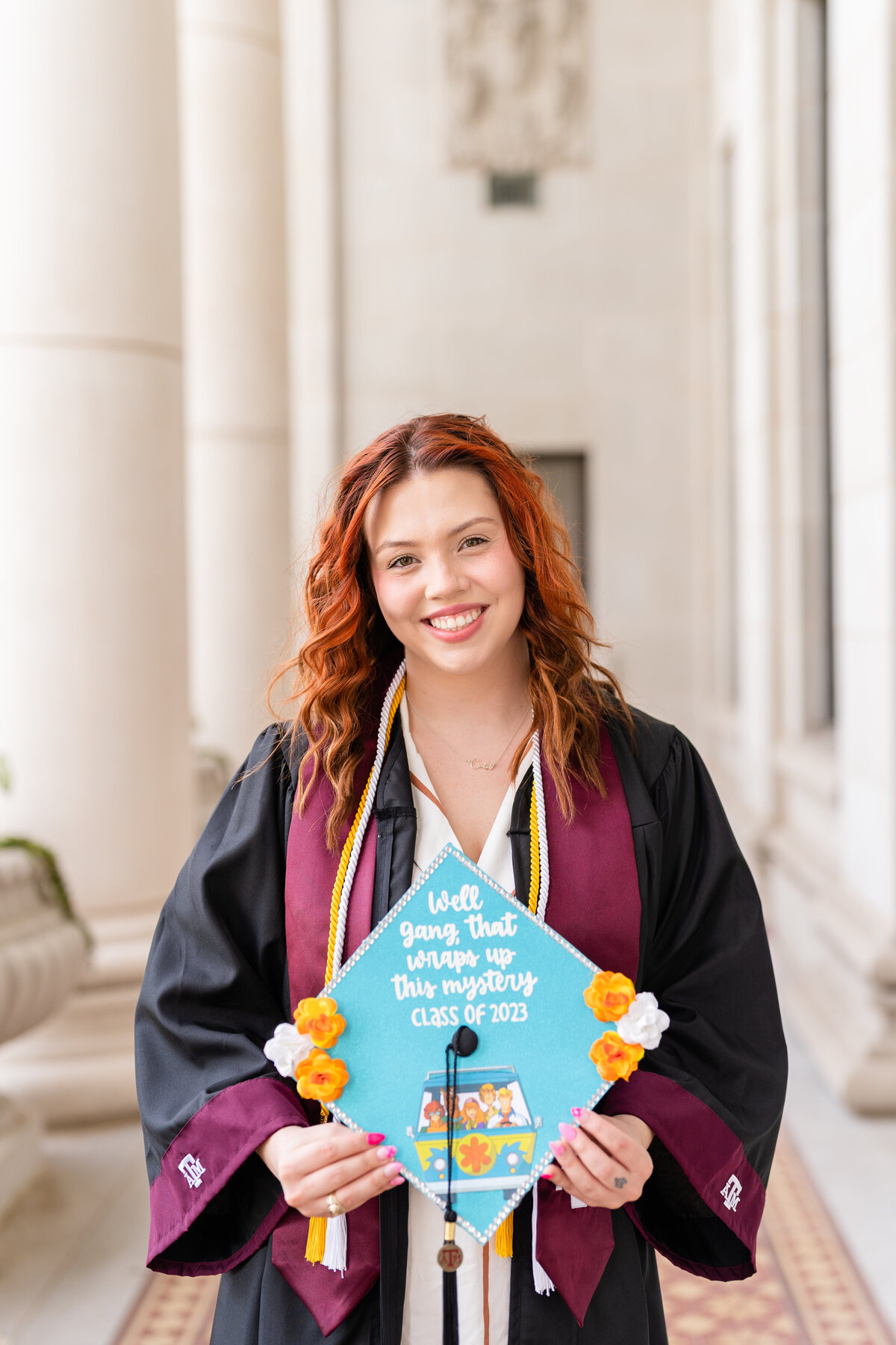 Texas A&M senior girl smiling and holding up decorated grad cap while wearing gown, Aggie stole and cords in the columns of the Administration Building