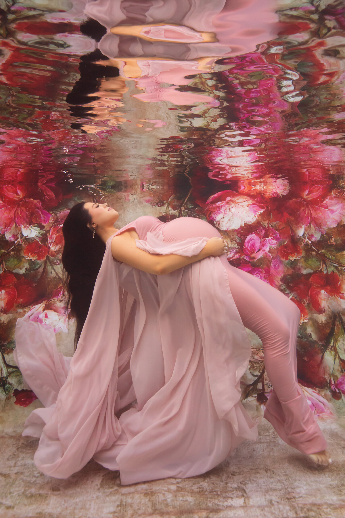 A woman in a flowing pink gown floating underwater against a vibrant floral backdrop of pink and red blossoms. Her serene expression and graceful pose create a dreamy, ethereal feel, enhanced by the soft, flowing fabric enveloping her like petals in bloom.
