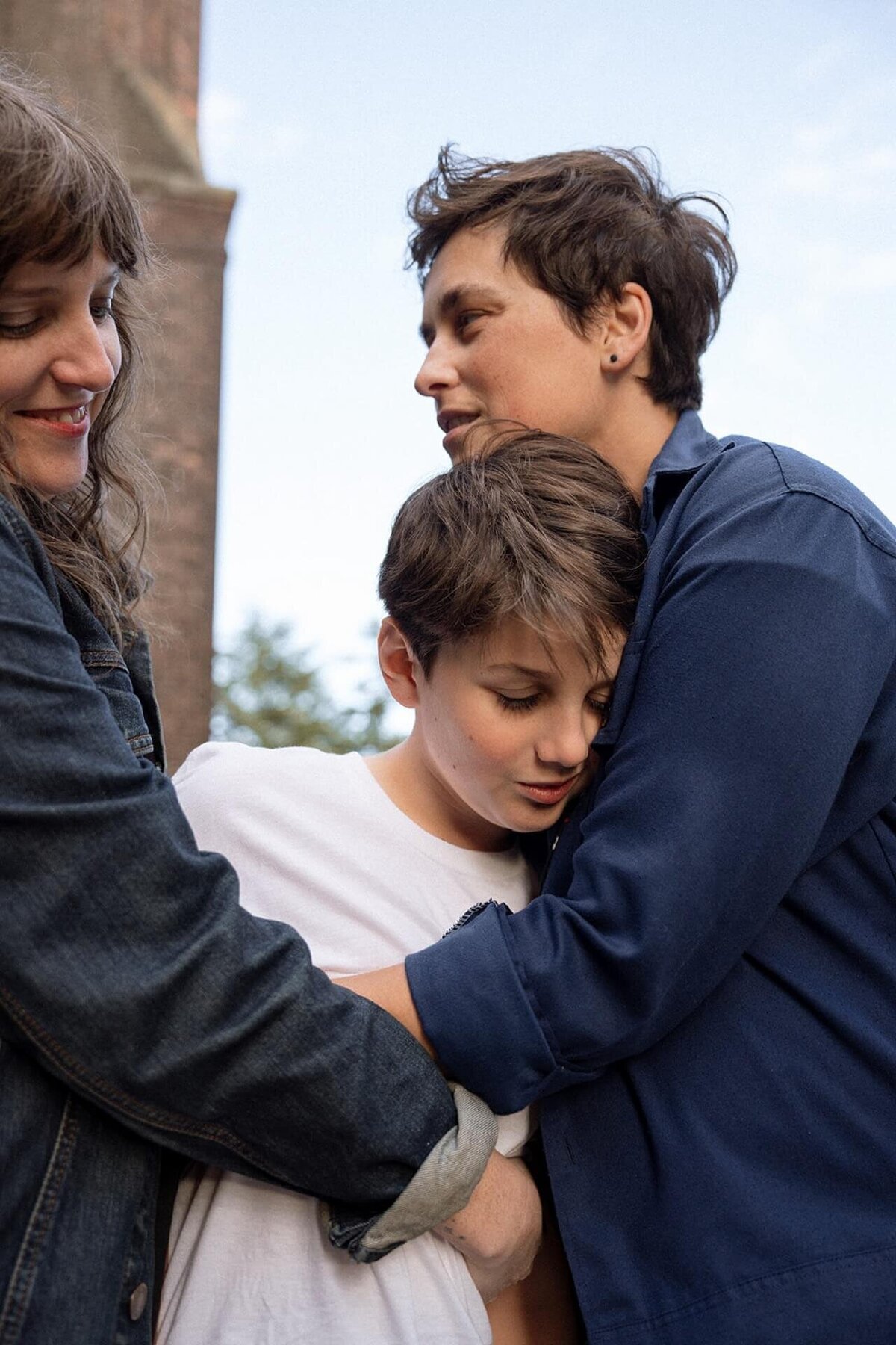 two moms wearing dark colors hugging their teenage son outdoors