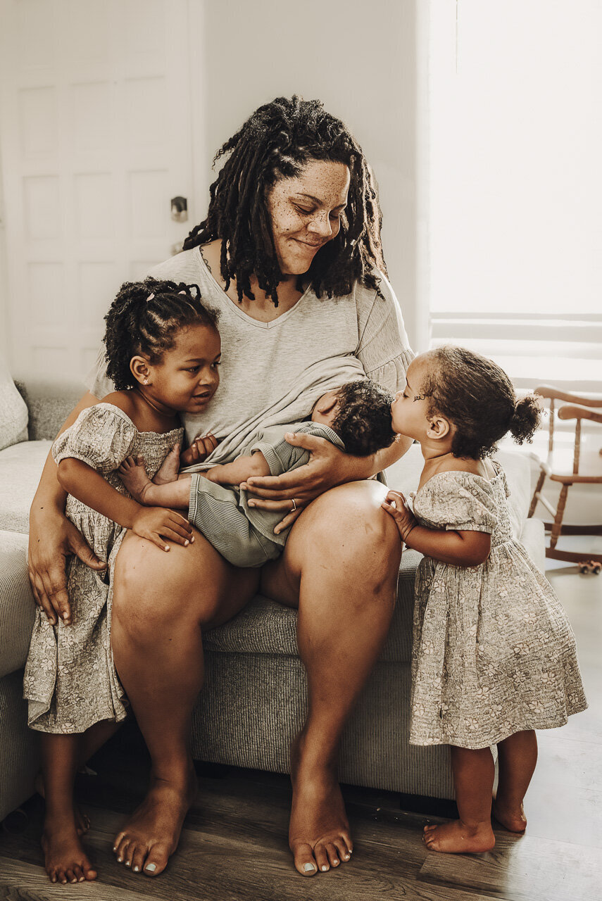 Sweet lifestyle newborn photography in Statesboro Georgia: mother nursing newborn as one sister kisses baby's head and other sister hugs Mom and looks at baby