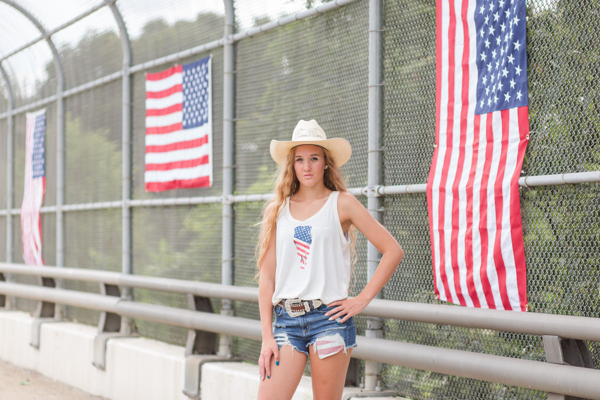 Waynedale High School girl standing on bridge decorated with flags for the fourth of July, she is standing and wearing blue jean shorts with red, white and blue pockets sticking out, along with a white tank top and a cowgirl hat.  Patriotic inspired photo, photographed by Jamie Lynette Photography Canton Ohio Photographer