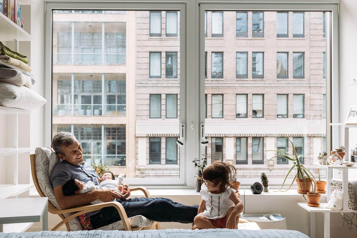 Newborn Photographer, dad sits with new baby as toddler sibling looks on in city apartment