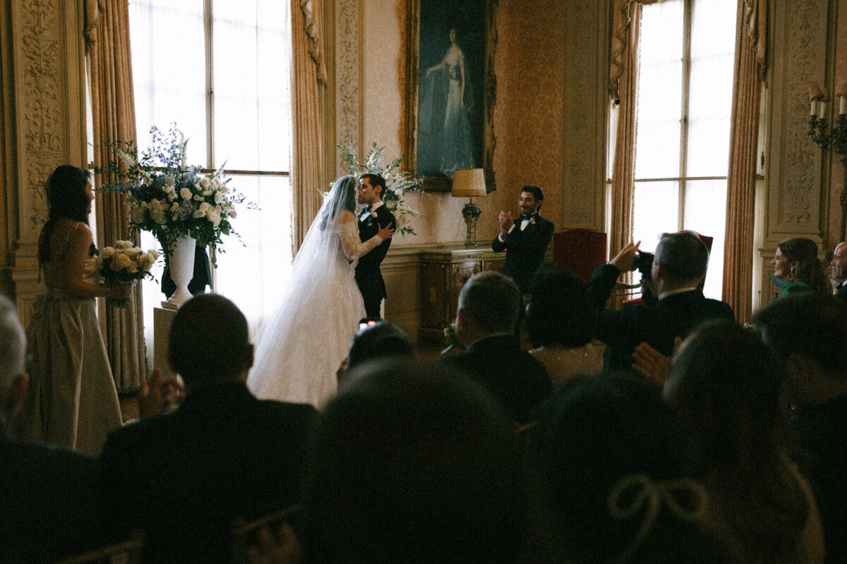 Bride and Groom having their first kiss inside the Rosecliff Mansion in Rhode Island while their friends and family celebrate, documented by Andrea-Marie Photography
