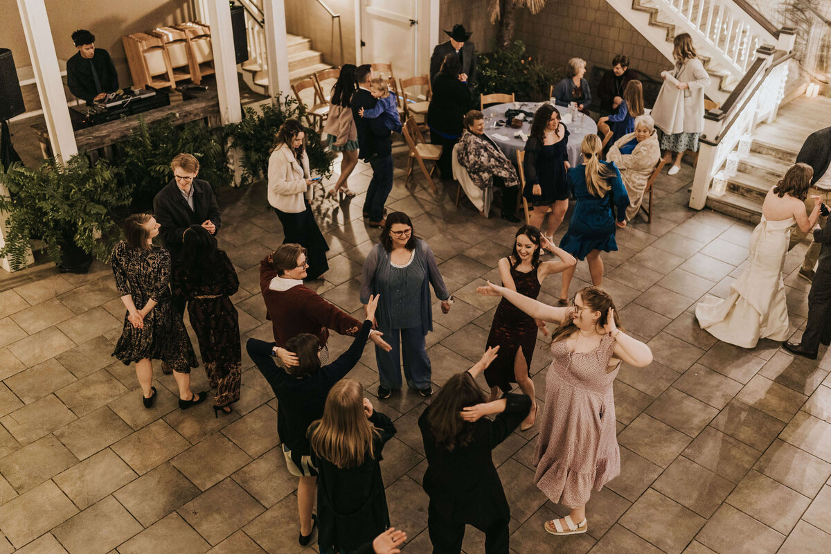 A group of people dressed in semi-formal attire dance and mingle at an indoor event, while others sit at tables or stand nearby. A DJ plays music in the corner, and a staircase is visible in the background.
