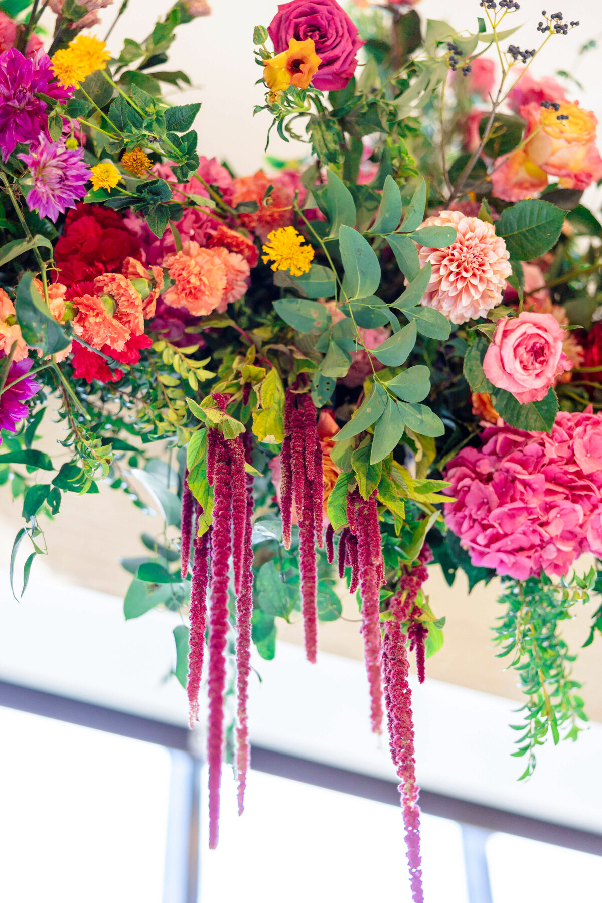 A vibrant floral arrangement featuring a mix of pink, red, and yellow flowers with lush green leaves and trailing amaranth blooms. The background shows a softly blurred window with natural light.