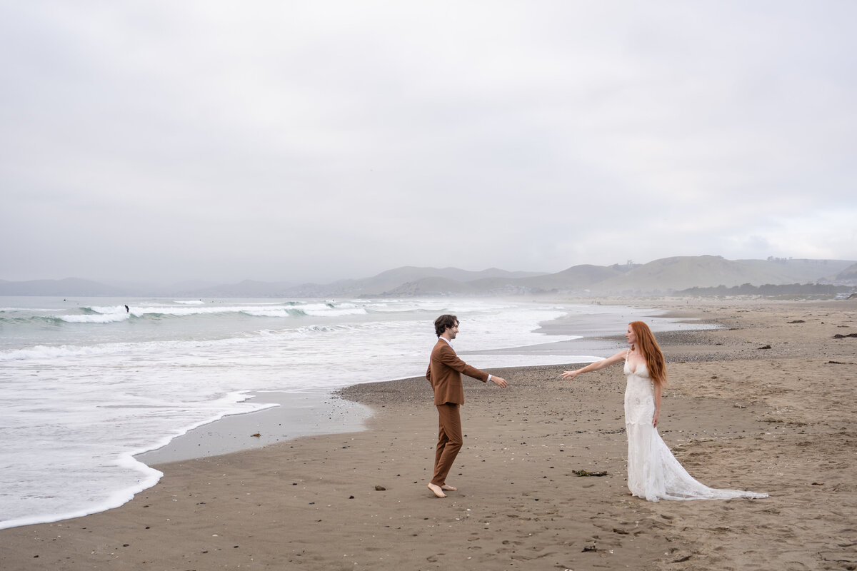 Bride and groom at the Morro Bay beach barefoot at golden hour reaching for each others hands in a lace Claire Pettibone gown with train