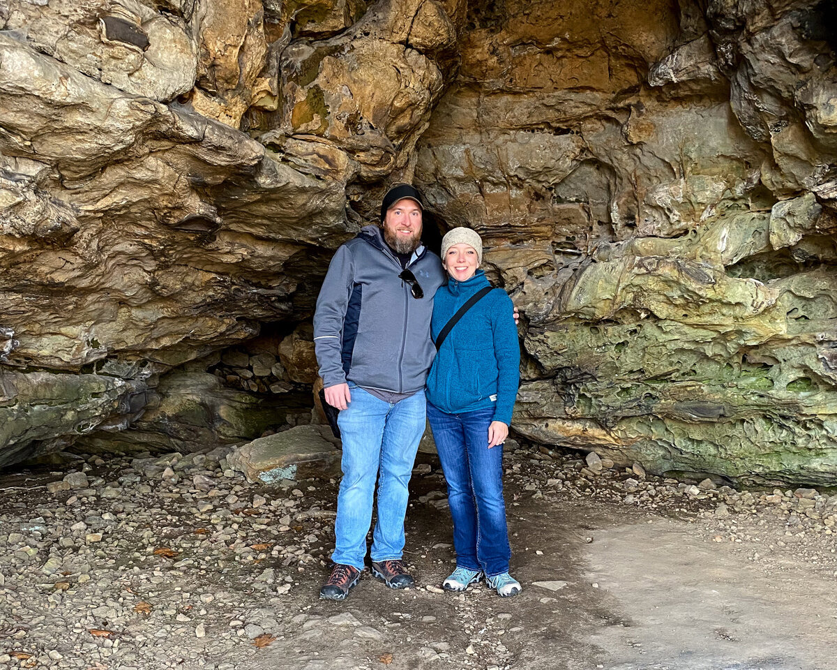 husband and wife standing on a rock formation, next to each with arms around one another, looking at the camera smiling, wearing jackets, jeans, hiking boots, and ear warmers