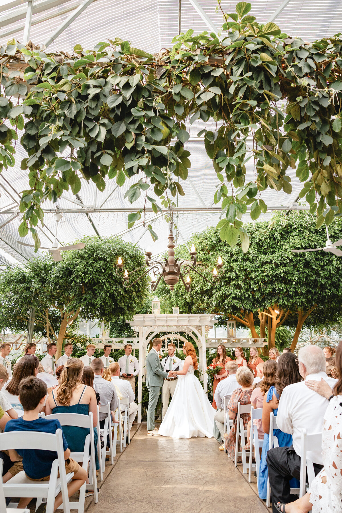 The bride and groom stand at the altar inside a greenhouse-style venue with a high ceiling covered in vines. Guests sit in white chairs, watching as the couple exchanges vows.