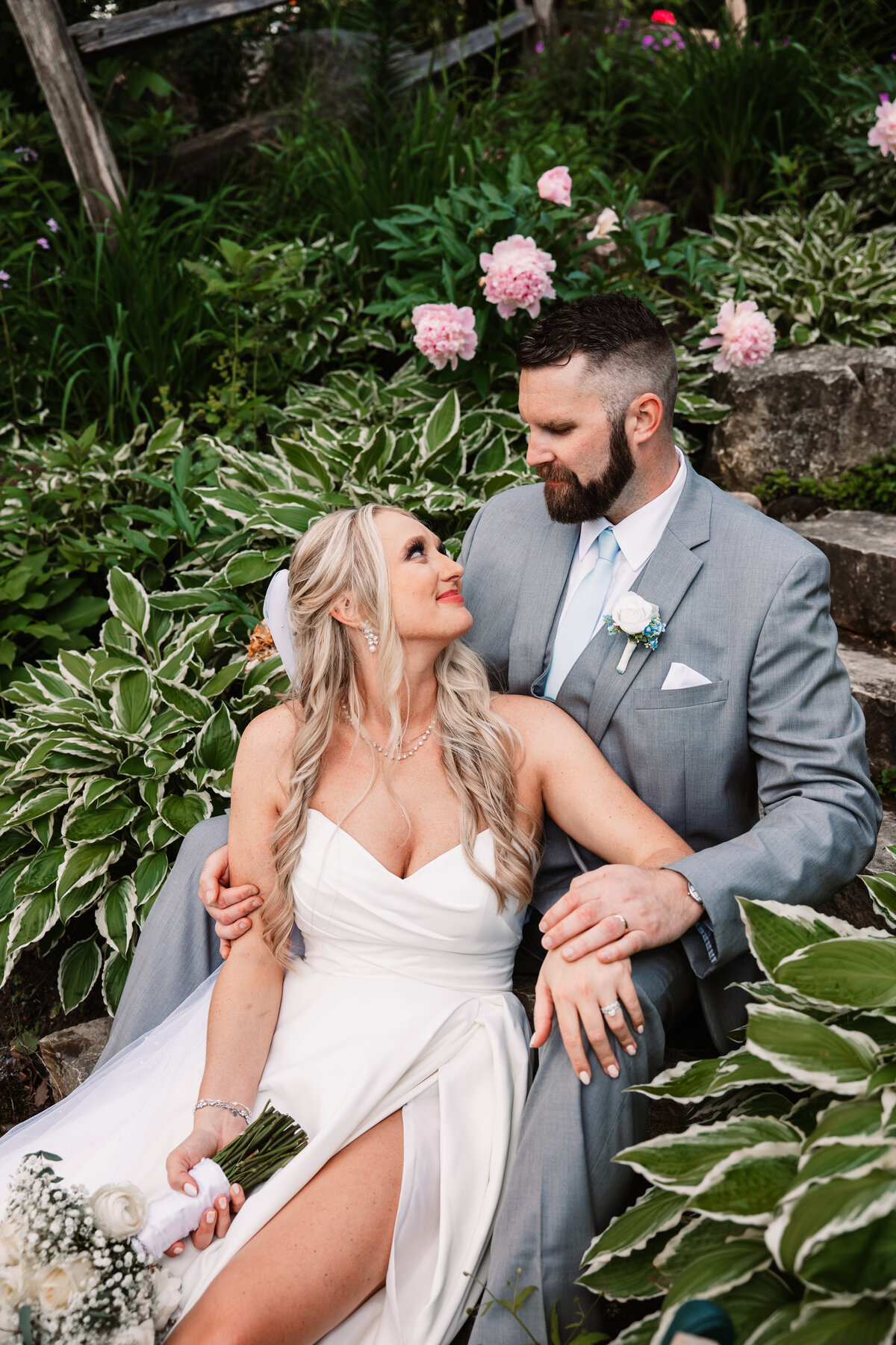A bride and groom sit on steps of stairs in a flowering garden cuddling and smiling at each other