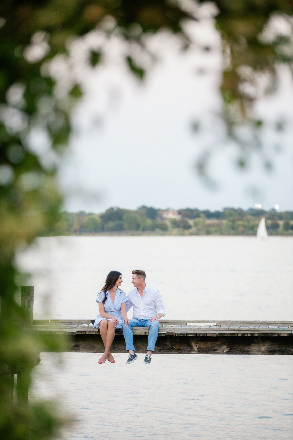 Engagement portrait of a couple sitting on a pier at White Rock Lake, Dallas, with a sailboat far behind them.