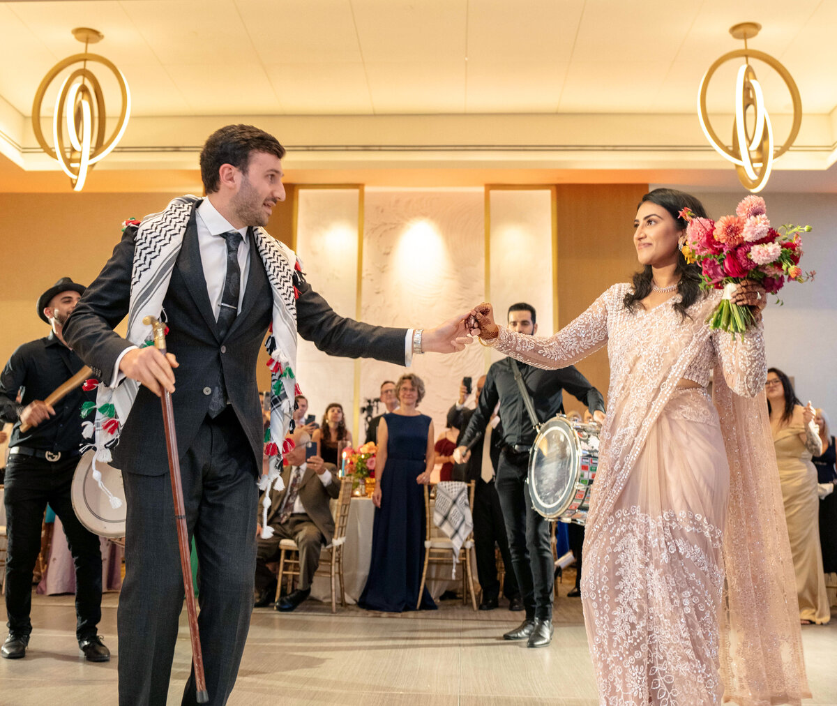 A couple dressed in formal attire dance at a celebration. The man wears a keffiyeh and holds a cane, while the woman in a decorative gown holds a bouquet. Musicians with drums perform in the background. The setting is a decorated event hall.