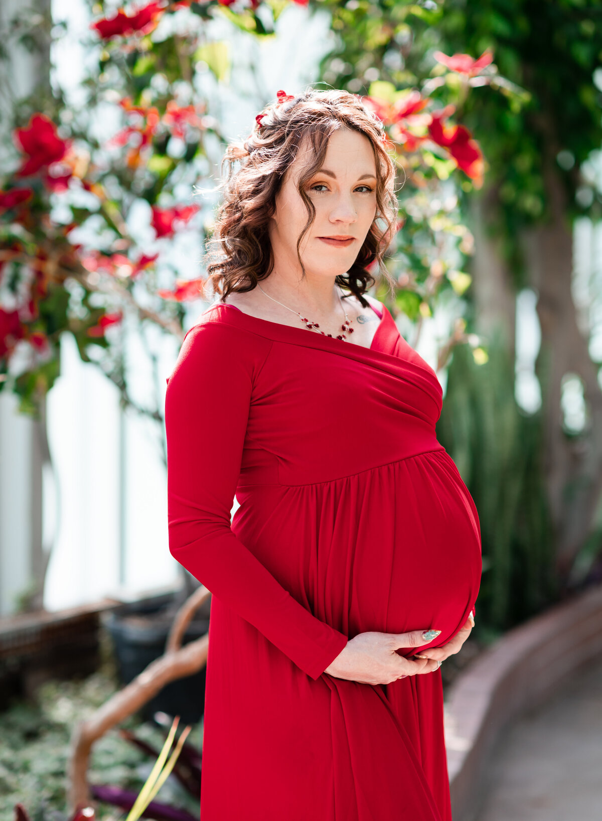 A radiant expectant mother in a red gown poses gracefully in a sunlit garden