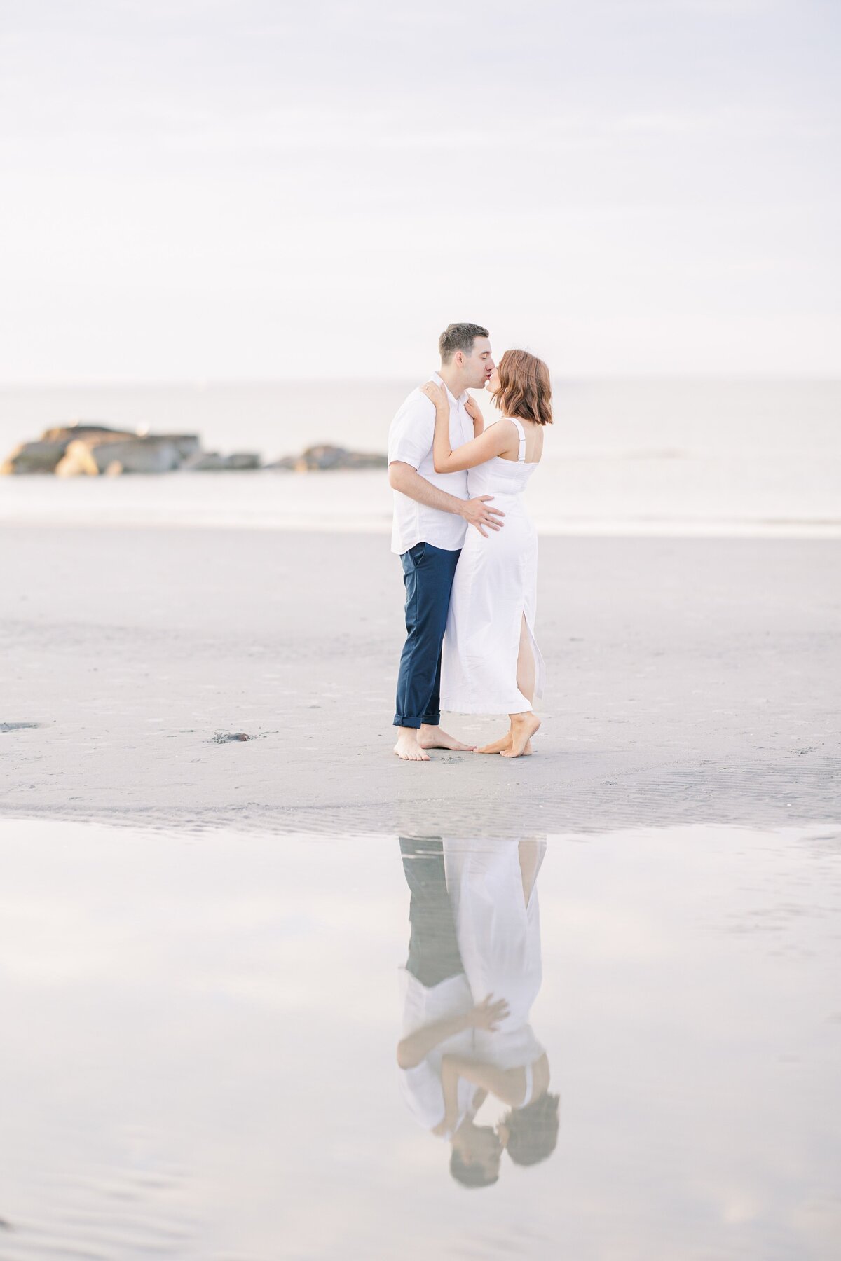 A couple kissing with their reflections mirrored in a puddle on the beach during their Wingaersheek Beach engagement session.