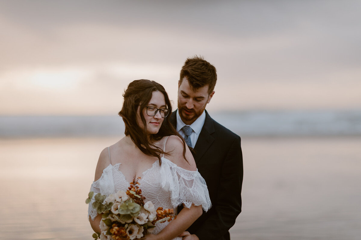 A bride leans into the chest of her groom while holding her bouquet and standing on a beach at sunset