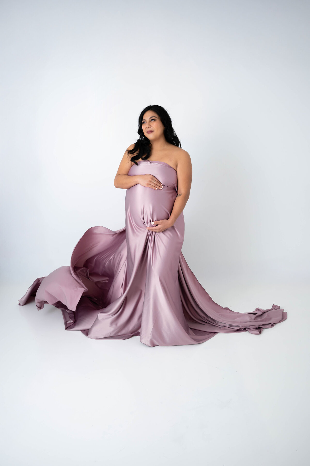 Expectant mother dressed in a flowy mauve silky fabric with a gentle smile looking off to the side. 