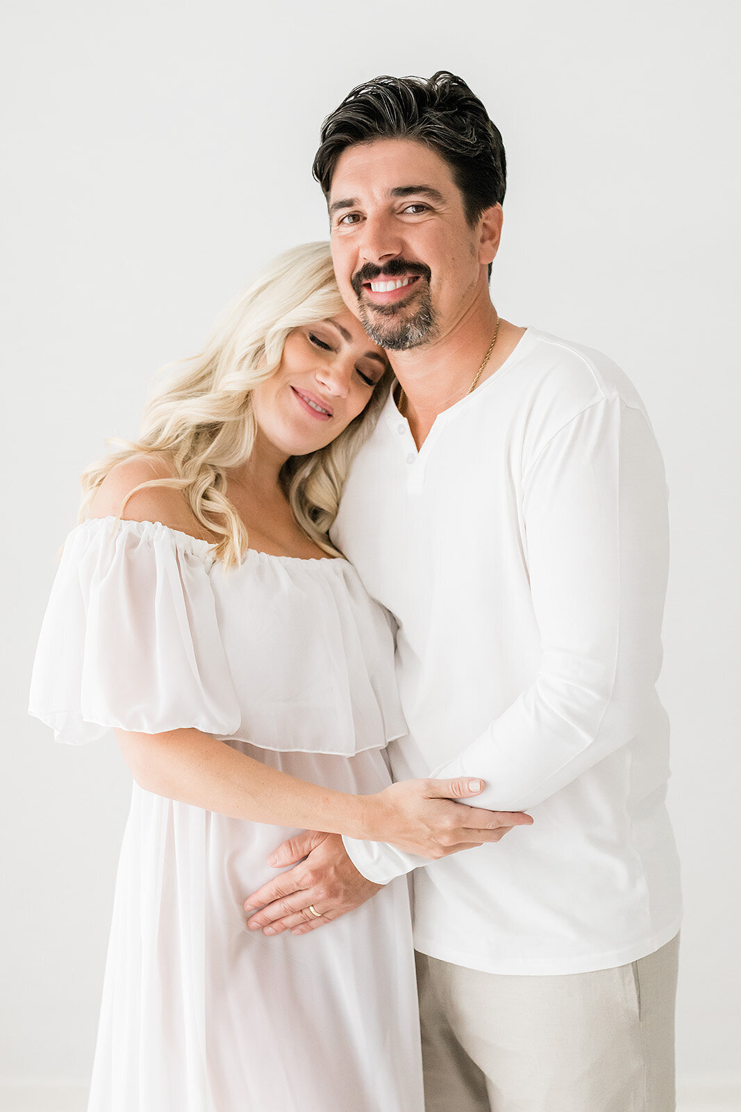 A couple stands together, smiling under the serene skies of Destin. The woman leans her head on the man's shoulder, both wearing white clothing, capturing a moment of warmth in this exquisite maternity photography by Andrea Krey.