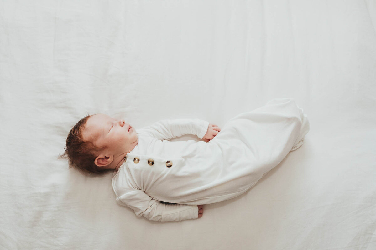 a newborn baby dressed in a white outfit lays on a white bed sleeping