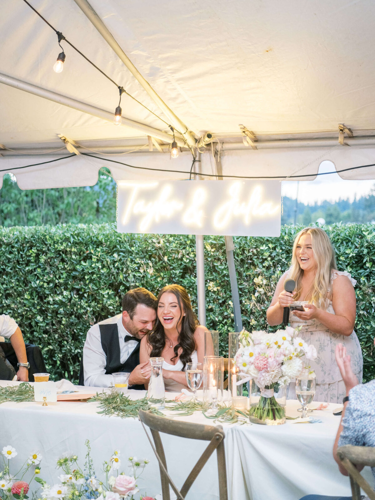 A bride's maid gives a speech at an outdoor wedding reception while the bride and groom embrace and laugh.