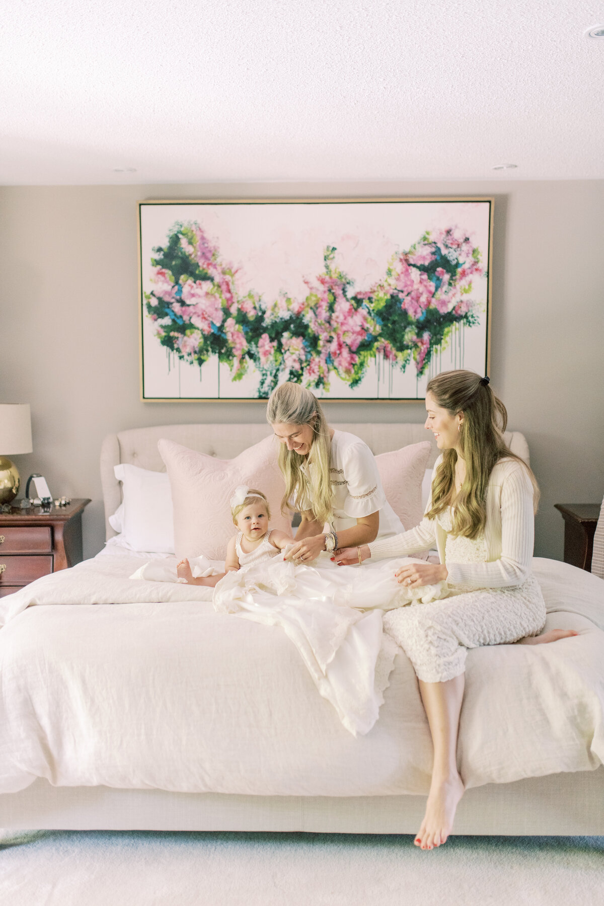 Serene portrait of mother and godmother cradling baby on soft bedding, mirror reflecting floral tones.