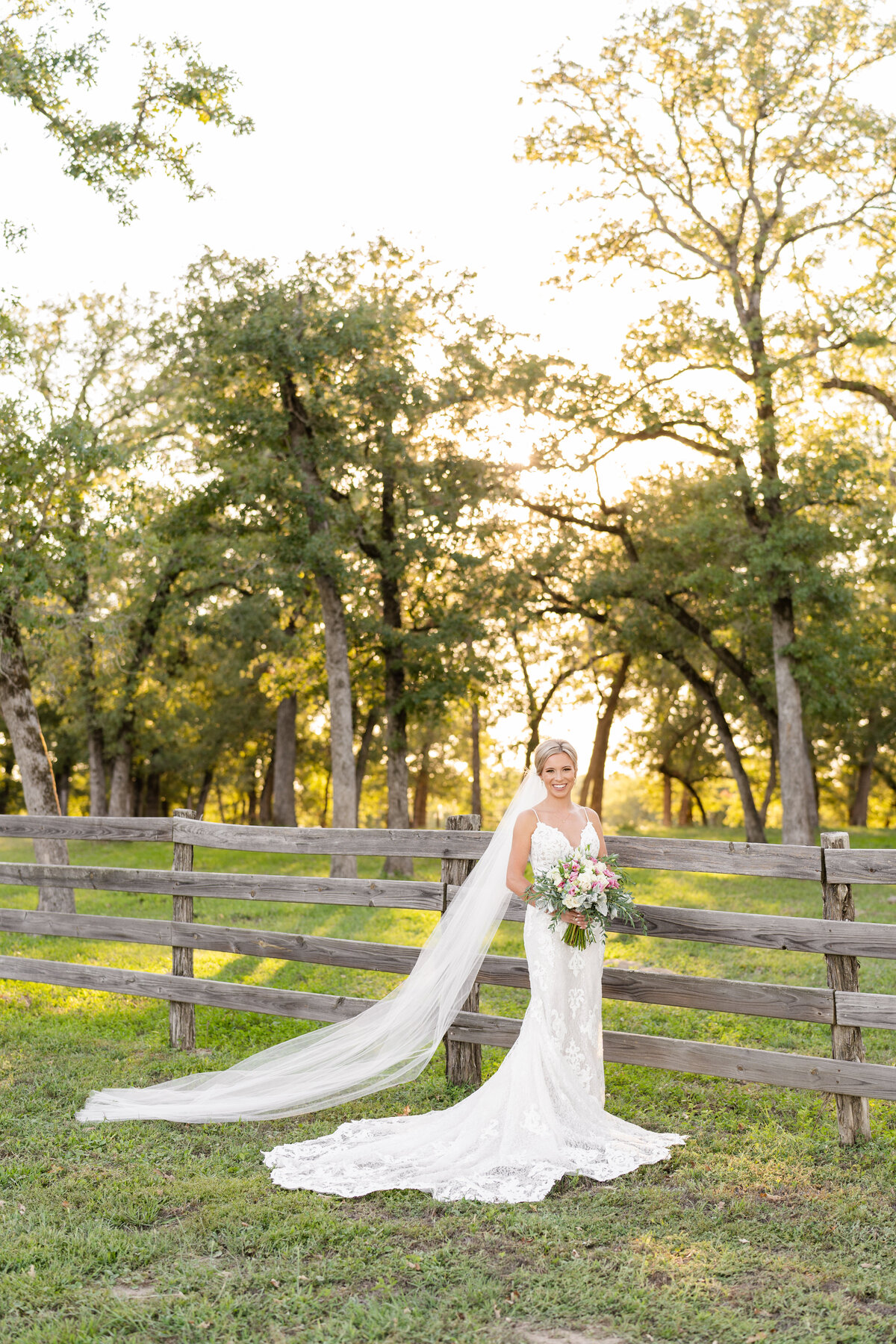 Bride holding bouquet with cathedral veil and smiling at camera next to a wooden fence and tall trees at sunset