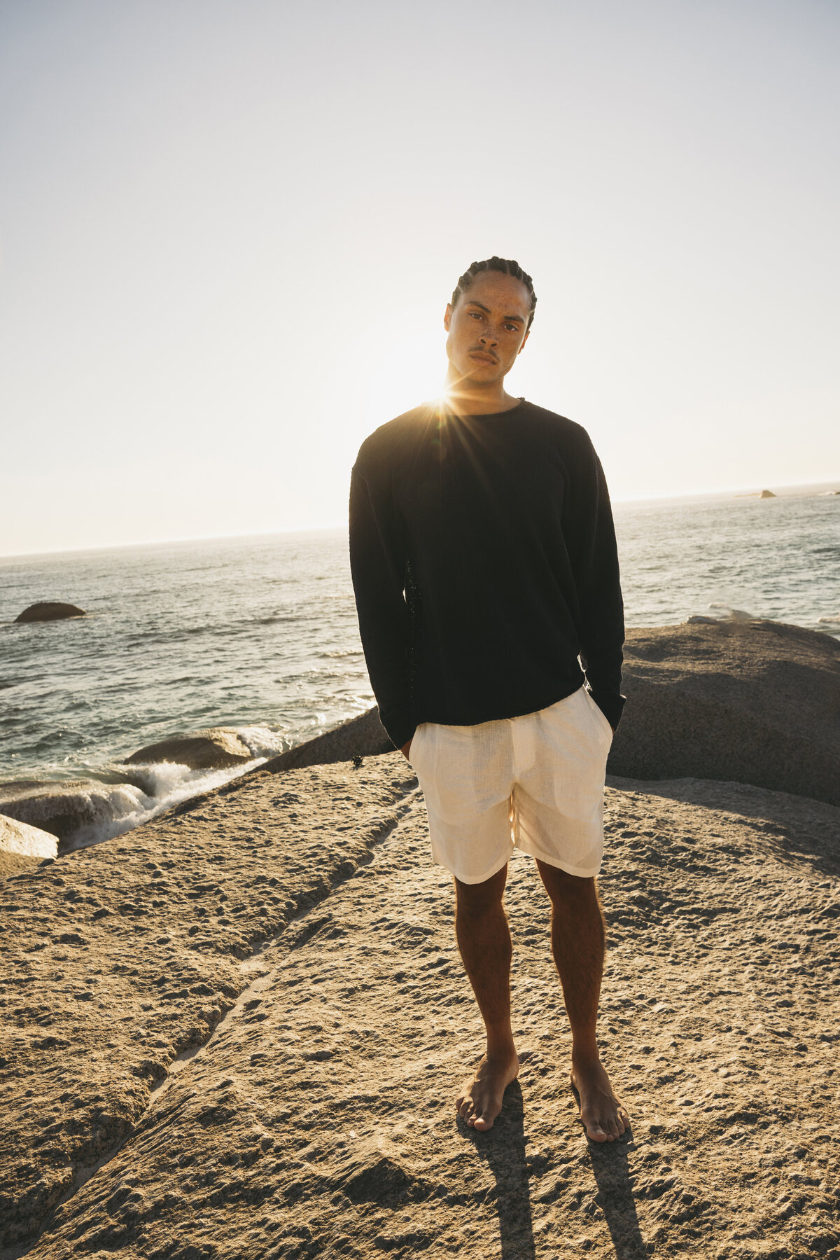 Full-body portrait of Timothe standing barefoot on a rocky coastline in Cape Town, with the sun setting behind him and casting a warm glow over the ocean.