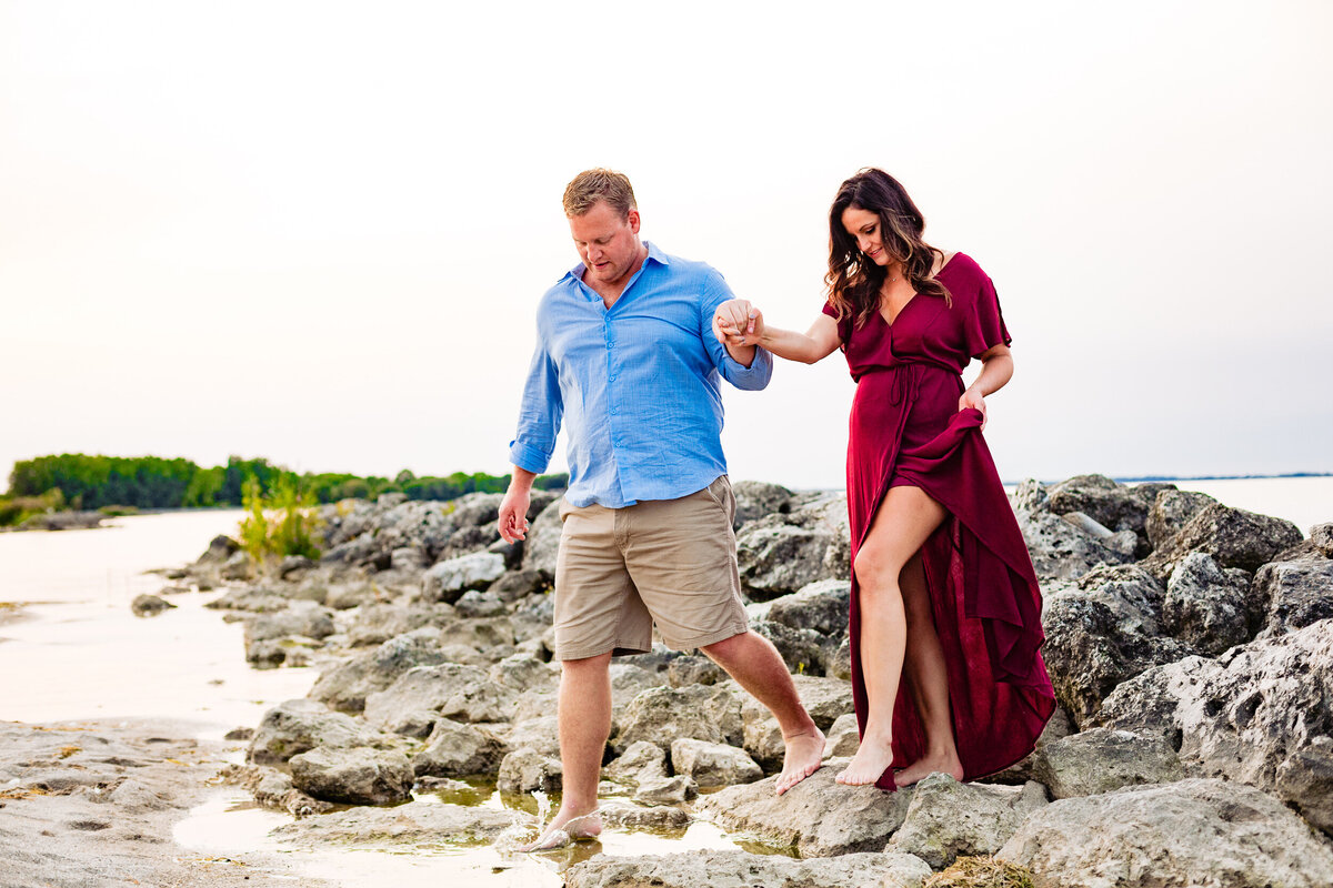 A couple walks barefoot hand and hand at Maumee Bay Resort. The bride wore a wine colored dress during their engagement photos.  Photo By Adore Wedding Photography. Toledo Wedding Photographers