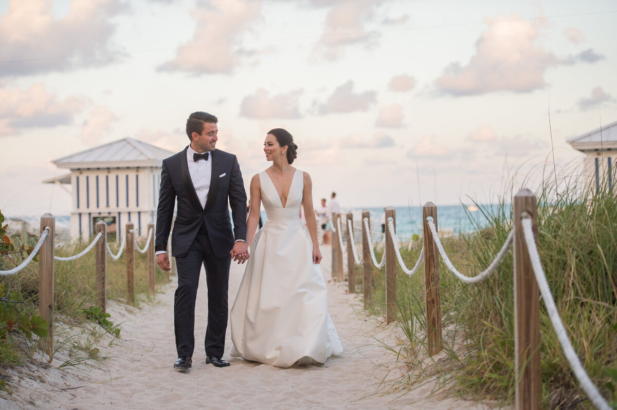 A bride and groom hold hands while walking down a sandy beach pathway, captured by Rodrigo Varela Photography.