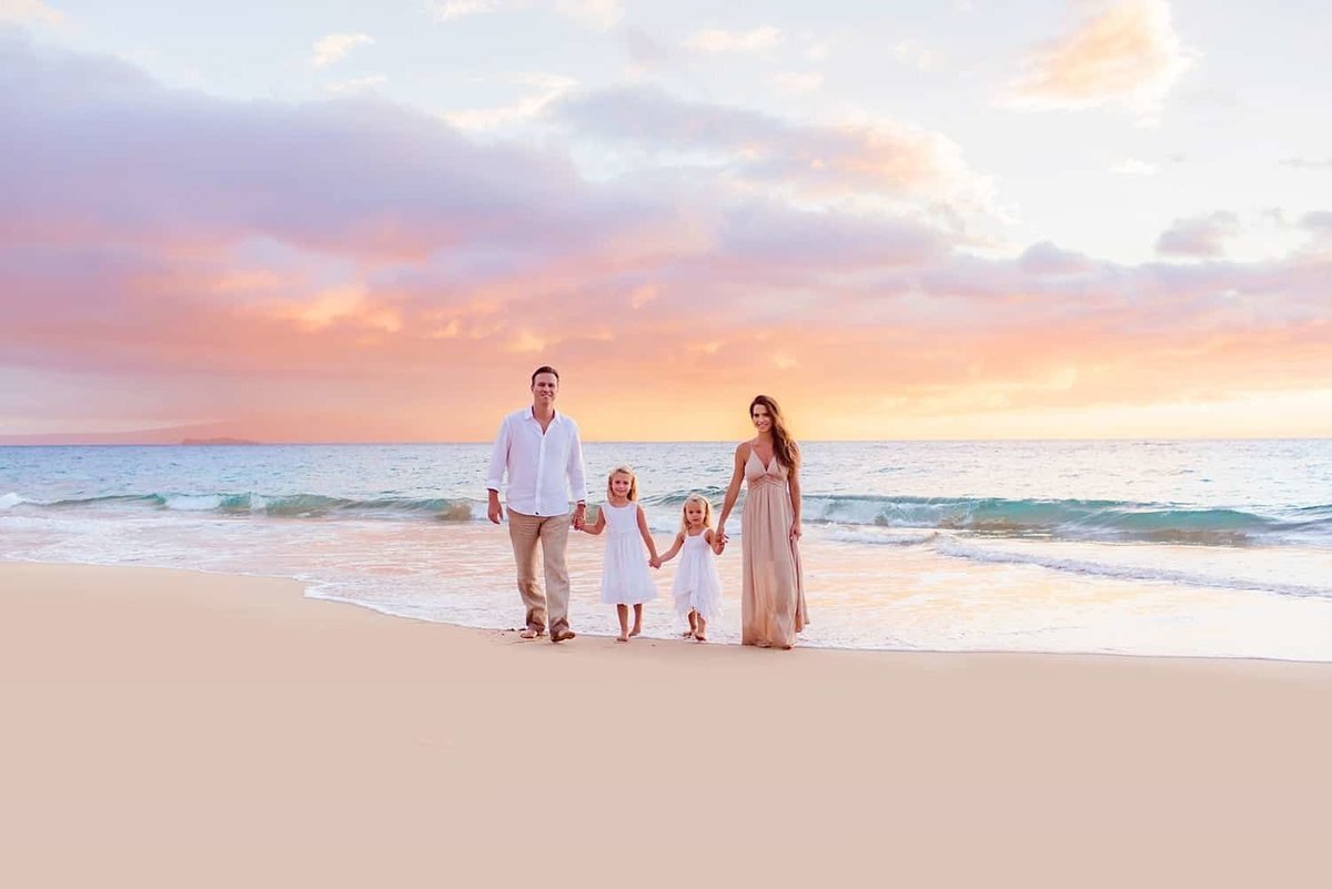Family portraits on the beach with Love + Water with everyone holding hands and walking toward the camera with pastel clouds in the background