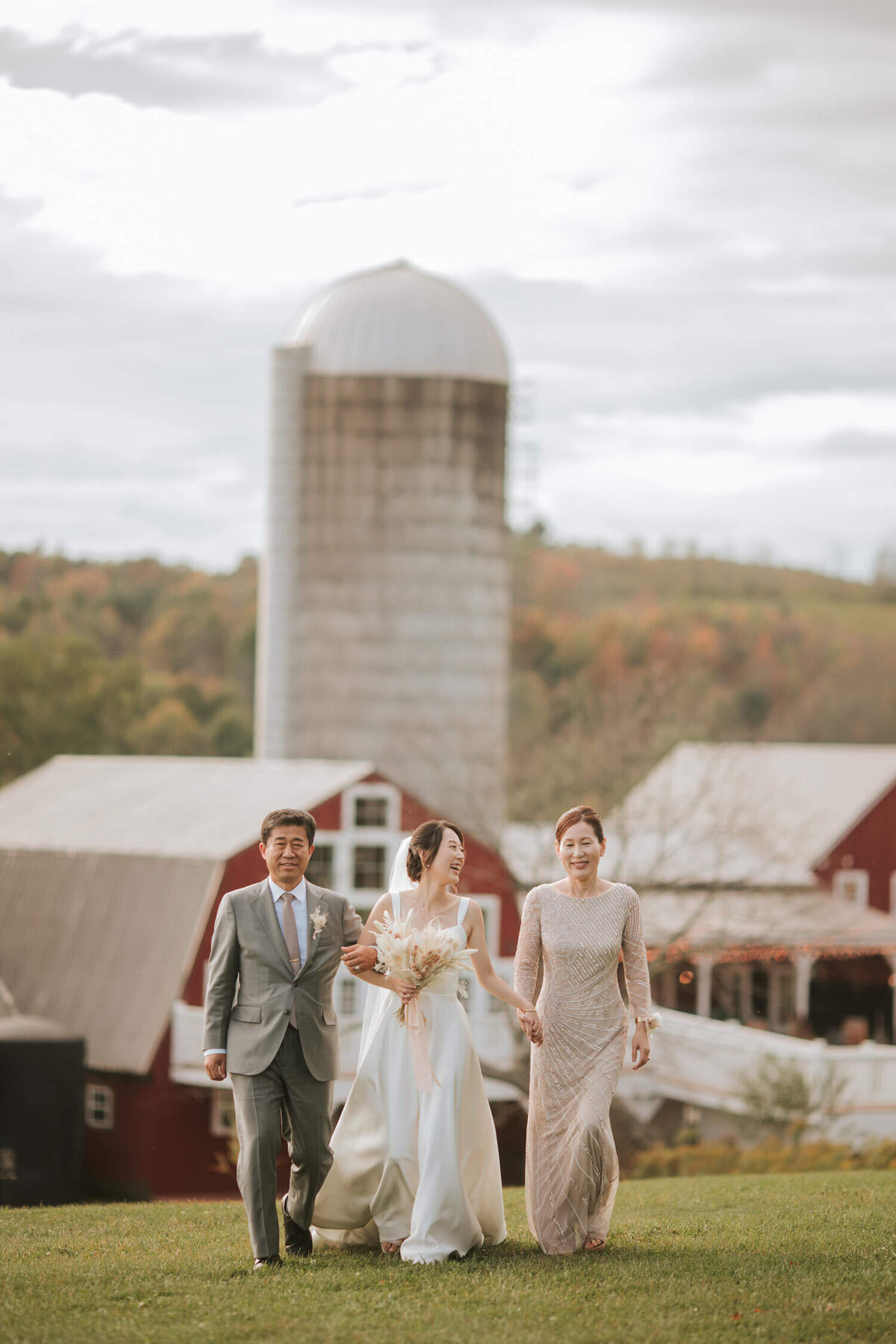 A bride walks arm in arm with her parents, smiling in front of a rustic barn and silo.